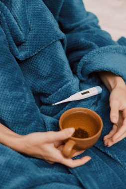 Mavi battaniyeye sarılmış bir insan, elinde termometreyle çay bardağıyla yatakta oturuyor..