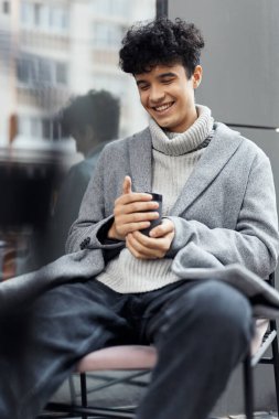 Sandalyede oturan, elinde cep telefonu olan ve kameraya gülümseyen genç bir adam. şık bir adamın moda portresi..