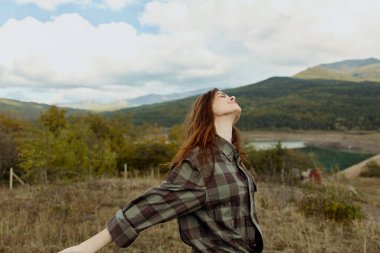 Huzurlu bir tarlada duran ekose gömlekli bir kadın. Arka planda görkemli dağlar, doğayı kucaklayan ve seyahat eden.