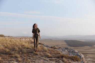 Tepenin tepesinde duran bir kadın, arka planda dağlarla doğa manzarası ve macera yolculuğu deneyimi.