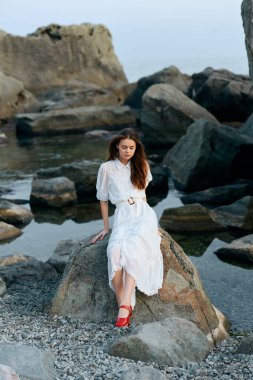 Beyaz elbiseli sakin bir kadın okyanus kayalarının üzerinde oturuyor. Etrafı kayalıklarla çevrili.