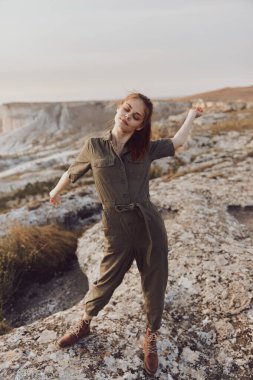 Zeytin rengi tulumlu genç bir kadın kollarını açık havada macera pozu vererek kayanın tepesinde duruyor.