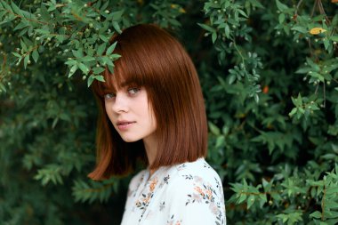 Kızıl saçlı kadın yemyeşil yeşilliklerin önünde duruyor, sırtı dönük.