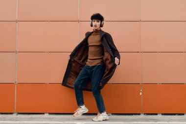 Moda. Kahverengi ceketli şık bir genç adam turuncu bir duvara karşı poz veriyor, elleri cebinde ve kafası yana yatık..