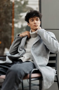 Gri bir ceket, beyaz balıkçı yaka kazak ve bir binanın önündeki sandalyede oturan bir gencin moda portresi..