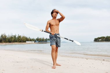 Aktif Sahil Macerası: Asyalı bir adam yaz güneşinde bir Paddleboard 'da egzersiz yapıyor ve sörf yapıyor, arkasında Parlak Mavi Okyanus ve Gökyüzü var