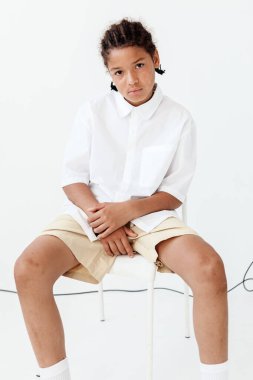 Beyaz gömlekli ve şortlu genç bir çocuk beyaz bir duvara yaslanmış modern minimalist bir sandalyede oturuyor..