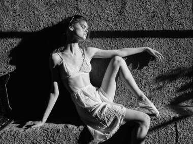 Dalgalı elbiseli zarif bir kadın güneşli bir pencerenin yanındaki taş duvarda oturuyor.