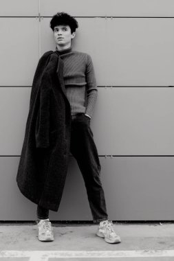 Süveter ve palto giymiş, elleri cebinde duvara yaslanmış genç bir adamın moda fotoğrafı. Görüntü siyah beyaz..