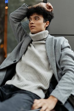 Gri bir kazak ve siyah pantolon giyen, elleri başının üstünde bir bankta oturan, kameraya bakan manken..