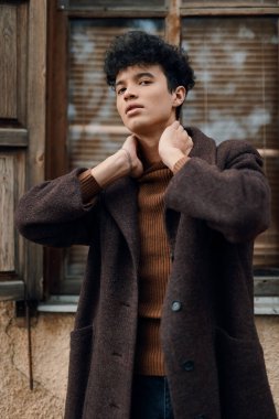 Kahverengi ceketli ve kaplumbağa yakalı şık bir genç adamın moda fotoğrafı pencereye yaslanmış, elleri boynunda ve kameraya bakıyor..