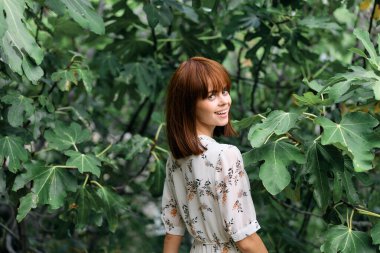 Çiçekli elbiseli güzel bir kadın çiçek bahçesinde yapraklı bir ağacın önünde poz veriyor.