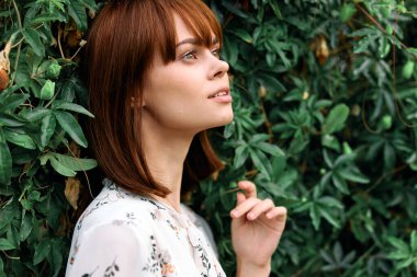 Canlı, kızıl saçlı bir kadın yeşil duvarın önünde duruyor. Yaprakları doğal bir şekilde ayarlanmış.