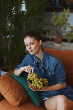 Önünde bir sepet üzümle kanepede oturan bir kadın.