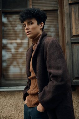 Kıvırcık saçlı, kahverengi ceketli, kaplumbağa yakalı kazaklı, ahşap panjurlu eski bir binanın önünde duran şık bir genç..