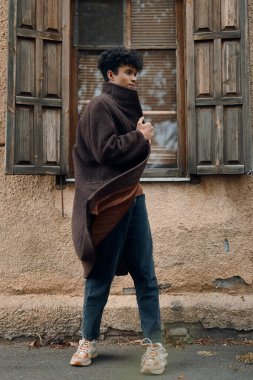 Kahverengi ceketli ve kot pantolonlu moda kadın eski binanın penceresinin önünde duruyor..