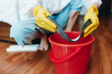 Lastik eldivenli bir adam bir kova suyun yanında yerleri paspaslıyor. Mavi gömlekli ve sarı eldivenli başka biri izliyor..