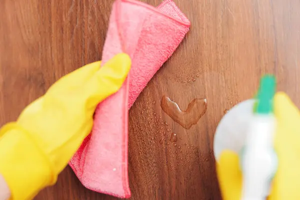 Stock image Woman cleaning wooden table with yellow rubber gloves and pink cloth, heart shape on surface.