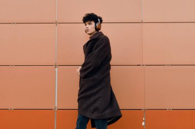 Paltolu ve eşarplı şık bir genç adam, turuncu bir duvara karşı kulaklıkla müzik dinliyor..