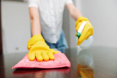 Sarı eldivenli bir kadın, bez parçası ve bir şişe temizlik çözeltisiyle masayı temizliyor. Bir kişi masa yüzeyini bir bezle siliyor ve temizlik ve hijyen için temizlik ürünü kullanıyor..