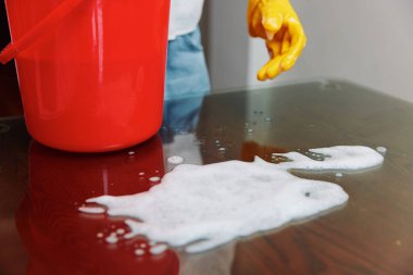 Yanında sabunlu su kovası ve kırmızı kovayla temizlik yapan bir kadın. Bir kadın bir kova sabunlu suyla masayı temizlerken, yakınlarda kırmızı bir kova görülüyor..