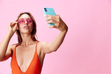 Şık bir mayo ve parlak pembe arka planda güneş gözlükleriyle selfie çeken şık bir kadın.