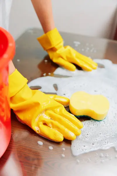 stock image Woman in yellow gloves cleaning a table with a sponge and soapy water. next to her, there is a red bucket.