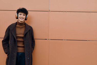 Kulaklık takan ve turuncu bir duvarın önünde gülümseyen genç bir adamın moda portresi..