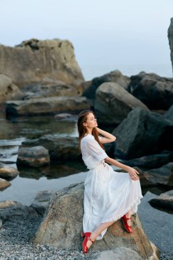 Beyaz elbiseli sakin bir kadın kıyı kayasında oturuyor, elinde kırmızı ayakkabılar, okyanus dalgalarına bakıyor.