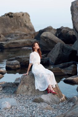 Uçan beyaz elbiseli bir kadın su kenarında kayanın üzerinde oturur, ayakları havada şakacıdır.