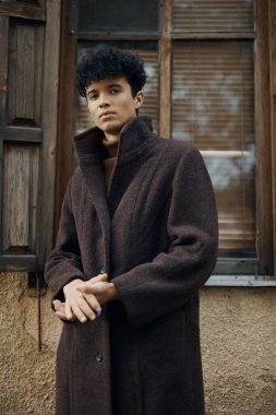 Moda: kahverengi paltolu bir adam elleri kalçasında ve başı yana eğilmiş bir şekilde pencerenin önünde duruyor..