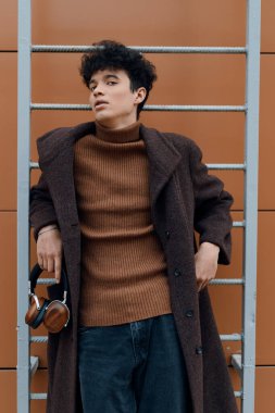 Kahverengi süveter ve palto giyen, duvara yaslanmış kulaklıkla müzik dinleyen modacı genç adam..