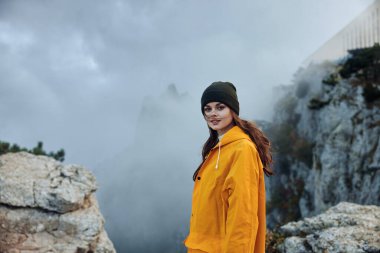 Sarı yağmurluklu ve bereli bir kadın görkemli bir dağ zirvesinde muzaffer bir şekilde duruyor.