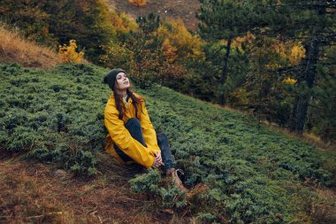 Doğada sakin bir an şık sarı bir ceket giyen bir kadın ormandaki bir tepede huzur içinde oturuyor.