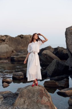 Beyaz elbiseli sakin bir kadın kayalık sahilde duruyor elleri saçlarında, okyanusa bakıyor.