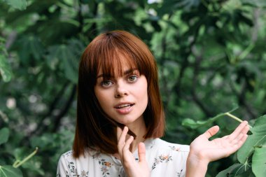 gür yeşil yaprakların önünde elleri üzerinde poz veren canlı kızıl saçlı kadın.