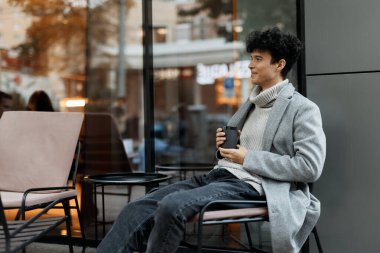 Şık bir binanın önünde oturan, elinde kahve fincanı ve elinde telefon tutan şık bir genç adam..