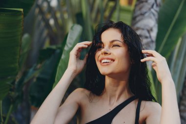 Güneşli bir günde, tropik palmiye ağaçlarının önünde poz veren siyah mayo giymiş mutlu kadın.