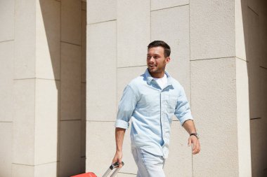 Kırmızı bavulu çeken, açık renkli bir binanın duvarına doğru yürüyen, güneşli bir günün keyfini çıkaran, gündelik kıyafetli mutlu genç adam..