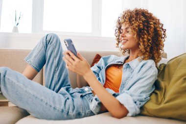 Evdeki rahat bir kanepede oturan mutlu bir kadın, elinde akıllı bir telefonla gülümseyen ve online oyunlardan zevk alan ve rahatça sohbet eden bir kadın..