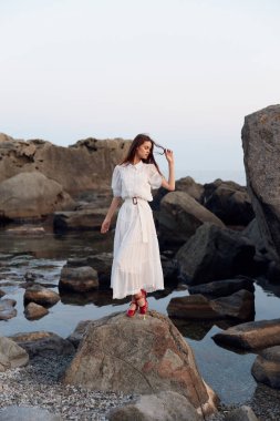 Beyaz elbiseli sakin bir kadın suyun kenarında duruyor elleri ceplerinde, güzelliği düşünüyor.