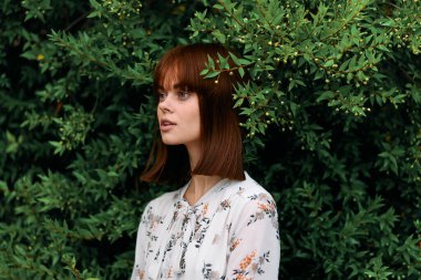 Canlı kızıl saçlı kadın, gür yeşil yapraklı çalıların önünde zarif bir şekilde duruyor.