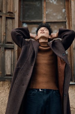 Kahverengi ceketli ve balıkçı yaka kazaklı genç bir adamın modadan portresi elleri kafasında ve gözleri kapalı bir binaya yaslanmış..