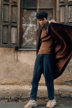 Kahverengi kazaklı ve mavi pantolonlu şık bir adam binanın önünde dikiliyor..