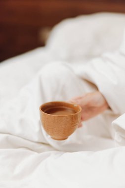 Yatakta oturan kişi elinde beyaz çarşafla bir fincan çay tutuyor..