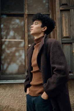 Kahverengi ceketli ve balıkçı yaka kazaklı şık bir genç adam pencerenin yanında duruyor, elleri cebinde gökyüzüne bakıyor..