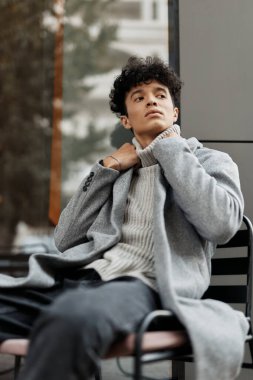 Gri montlu, elleri boynunda sandalyede oturan ve önündeki masada bir şeye bakan bir adamın moda görüntüsü..