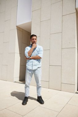 Açık renk kotlu genç adam düşünceli bir şekilde modern bir binaya karşı duruyor, güneşli bir günde gölge düşürüyor, tefekküre ve gündelik tarzına bürünüyor..