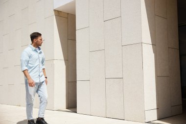 Günümüz minimalist binasının önünden geçen, derin düşüncelere dalmış sıradan kıyafetler giyen bir adam. Sahne, modern şehir yaşam tarzını ve yansıtıcı ruh halini yansıtıyor..