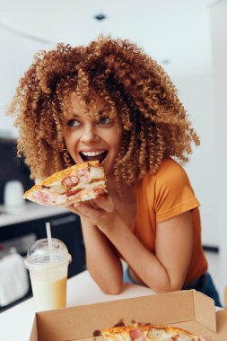 Kıvırcık saçlı kadın masada bir kutu lezzetli pizzanın önünde pizza yiyor.